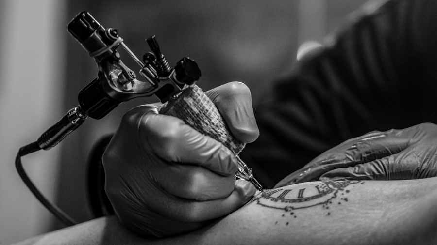 gloved hand holding tattoo pen to tattoo an arm with a clock tattoo